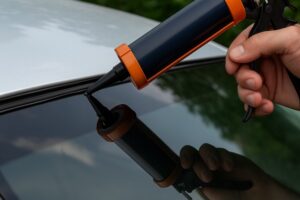 Close up of a person applying black automotive sealant to the edge of a car windshield using a caulking gun to fix a leaking windshield seal.